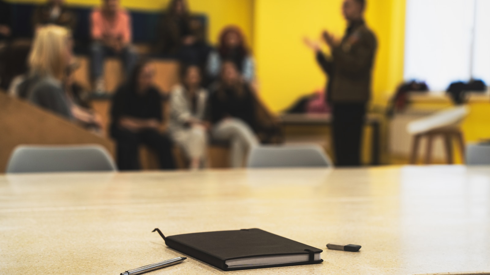 Notebook and pens on a table in front of a blurred meeting group and speaker